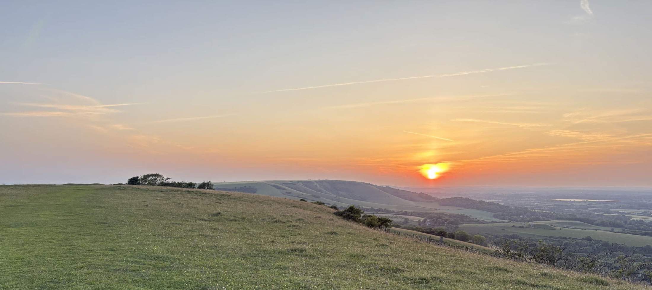 Sunset view over rolling green hills with a clear sky and the sun near the horizon.
